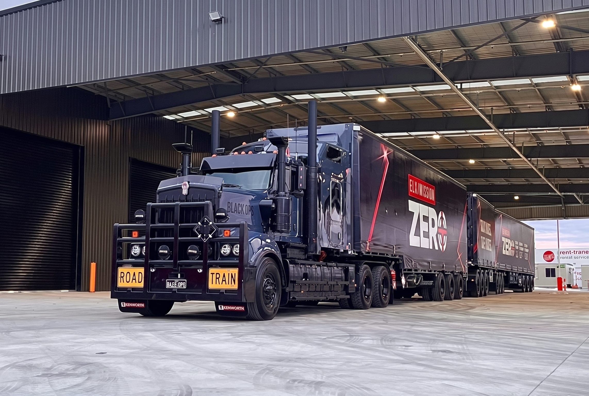 A black Kenworth road train with Mission Zero trailers parked in an open shed