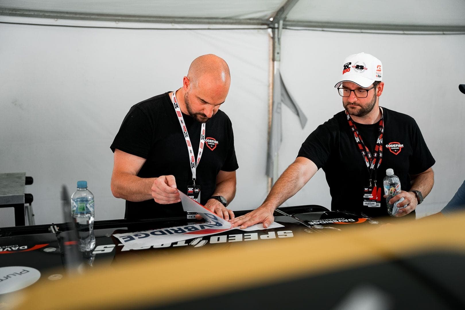 Two team members applying sponsor decals to race truck body panels in a pit marquee