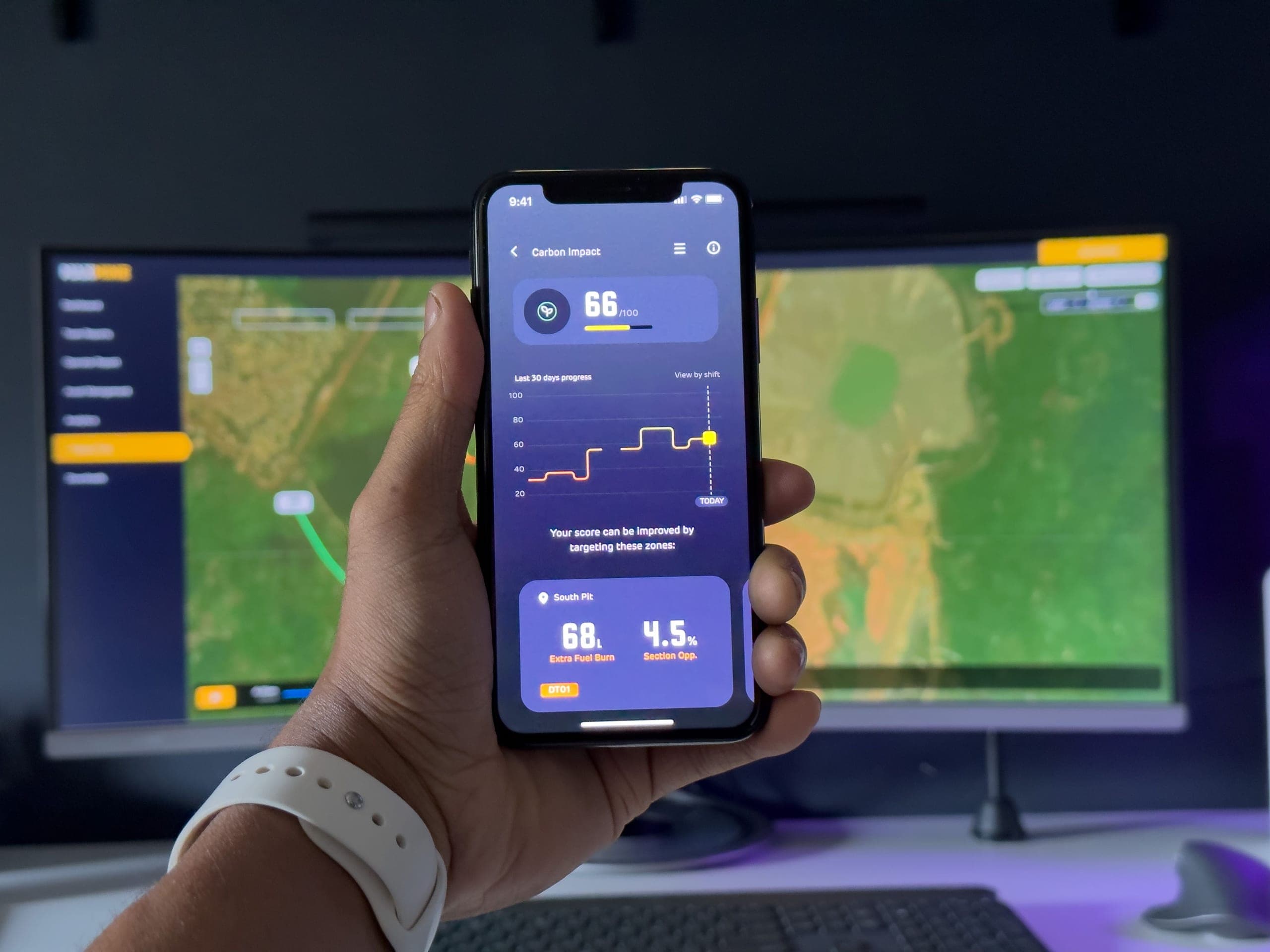 Hand holding a phone showing a mobile app with a score and chart, with maps on monitors behind