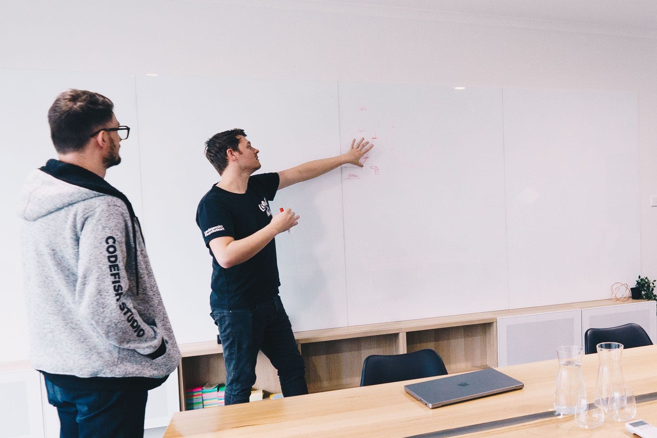 Two team members in a meeting room, one pointing at a whiteboard
