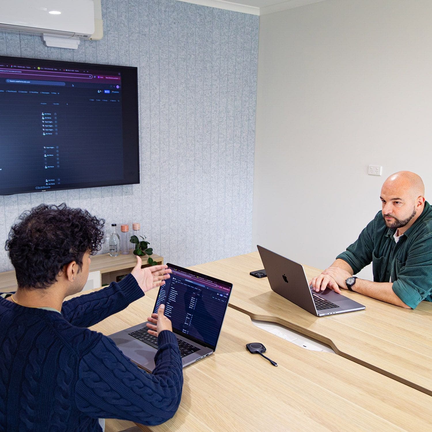 Two people discussing work on laptops in a meeting room with a screen on the wall