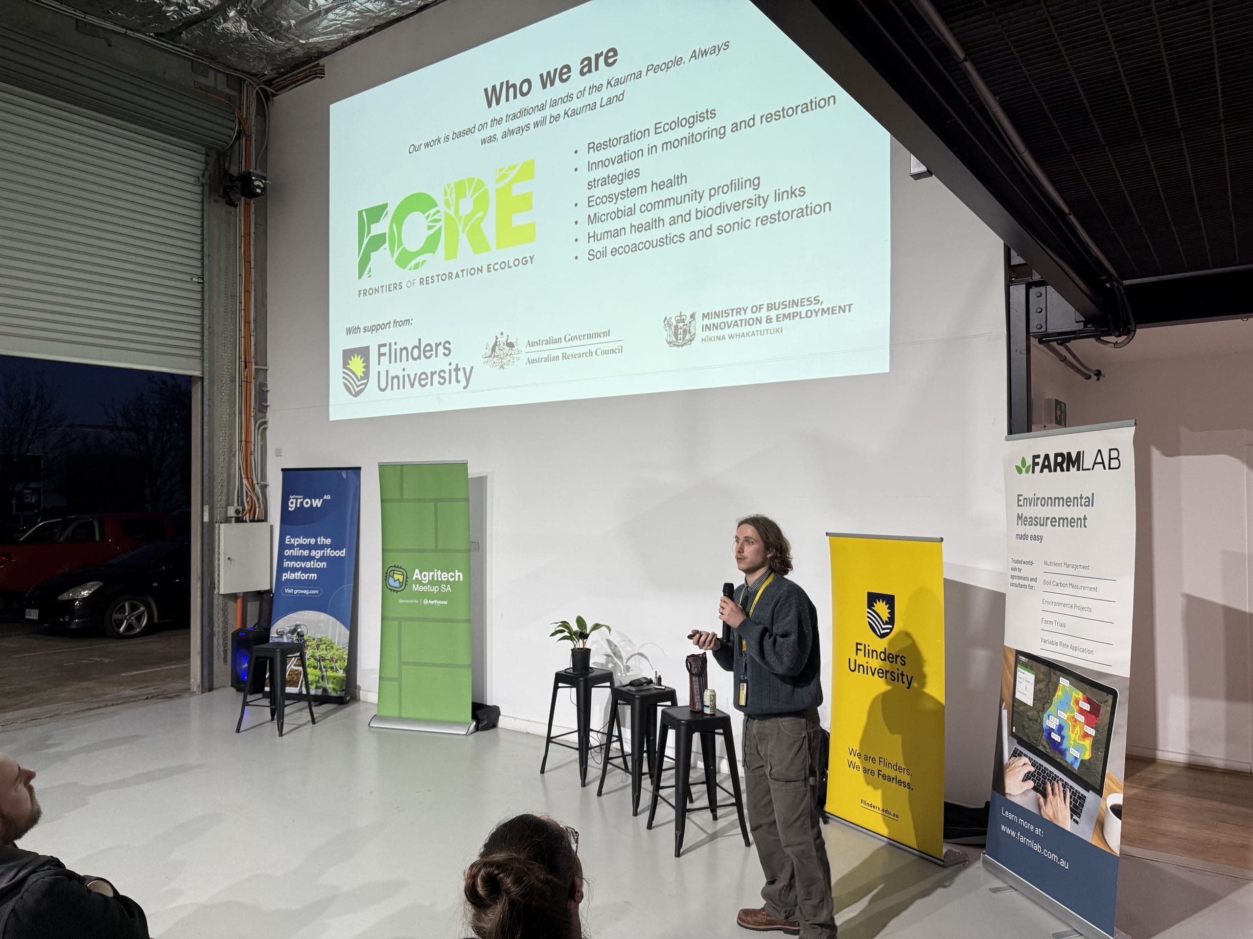 Speaker presenting at an agtech meetup in front of a projected slide and banners