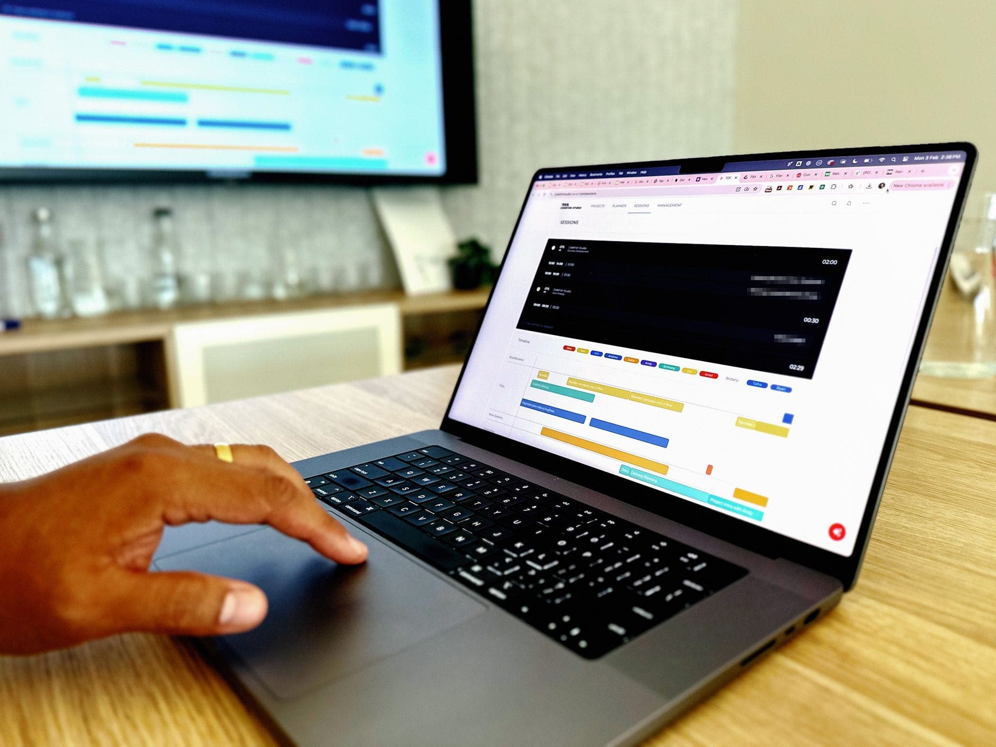 A hand on a laptop showing a colour-coded project Gantt schedule, with the same view on a wall screen