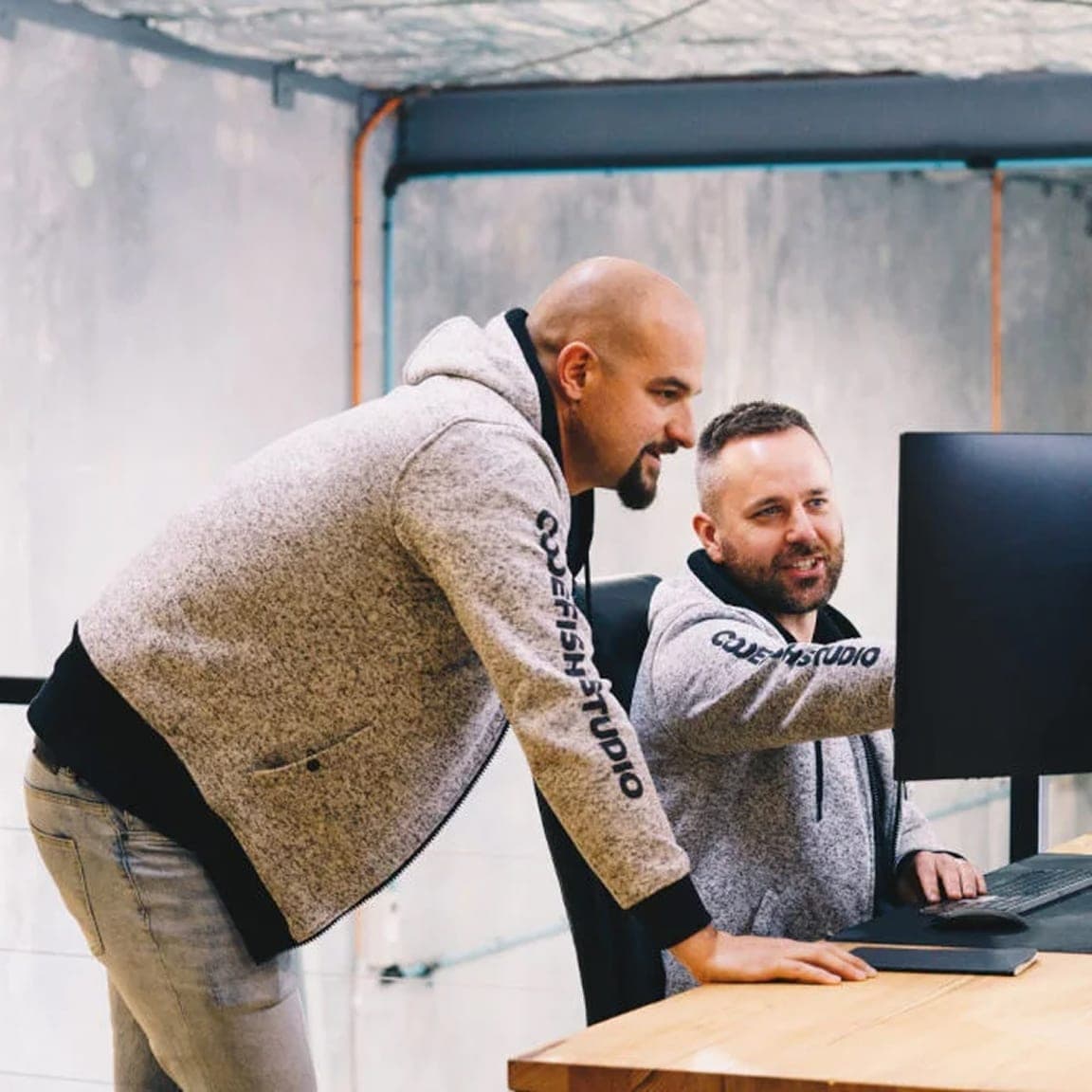 Two team members in branded hoodies reviewing work on a desktop monitor