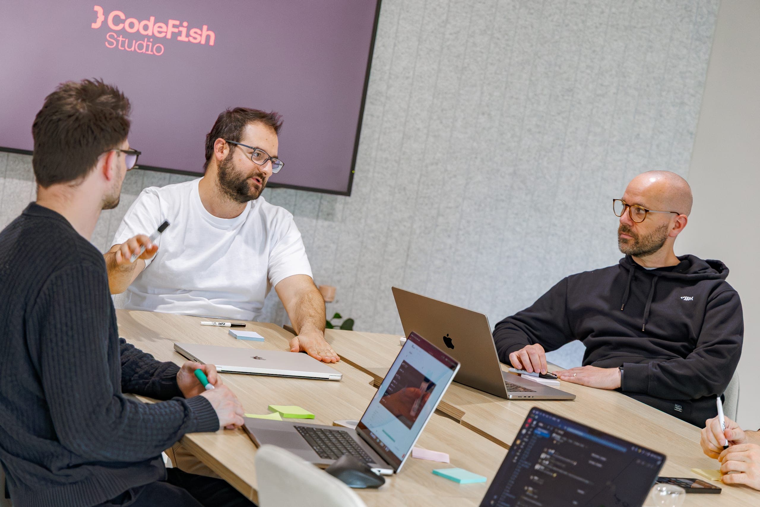 Three people talking around a meeting table with laptops and a screen behind them