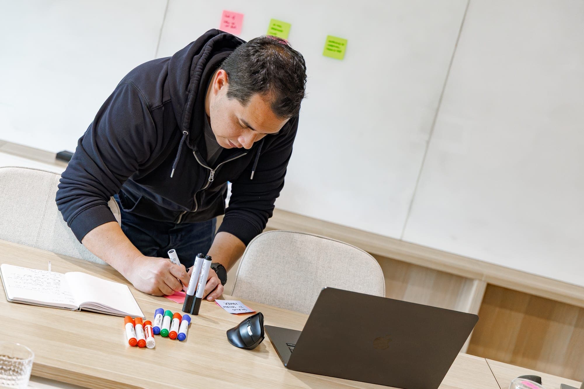 Person leaning over a table writing on a sticky note, with markers, notebook and laptop nearby
