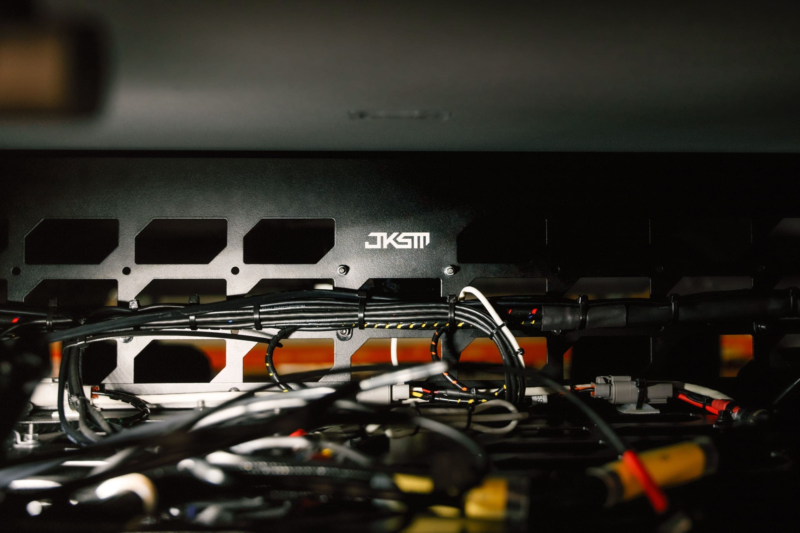 Close-up of a black perforated JKSM mounting panel above bundled vehicle wiring looms