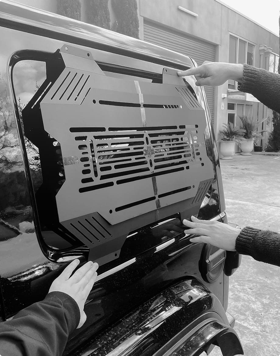 Two people holding a black storage panel in place over a vehicle rear window during a test fit